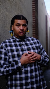 a young man in a plaid shirt leaning against a brick wall